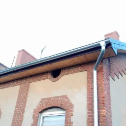 Metalowa rynna i obróbka blacharska na krawędzi dachu budynku z ceglaną elewacją i oknami z łukowatymi nadprożami.