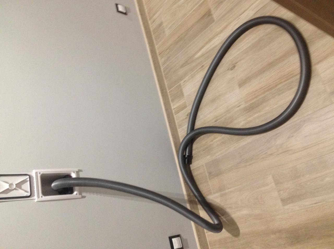 Szary, karbowany wąż podłączony do białej skrzynki w ścianie, leżący na podłodze imitującej drewno.