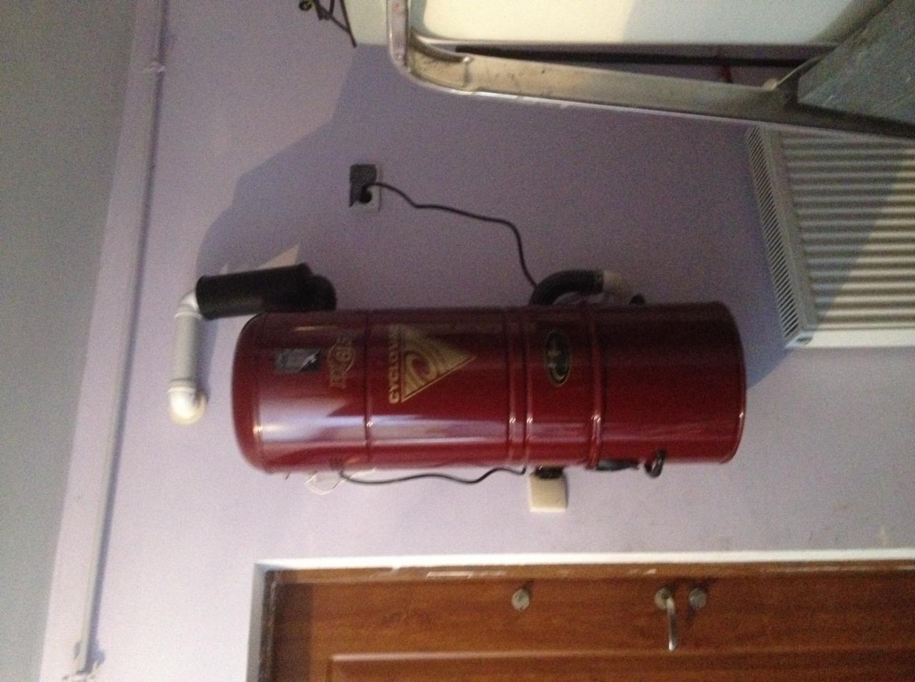 Czerwony, cylindryczny system centralnego odkurzania marki Cyclovac zamontowany na fioletowej ścianie, z widocznymi rurami i gniazdkiem elektrycznym.