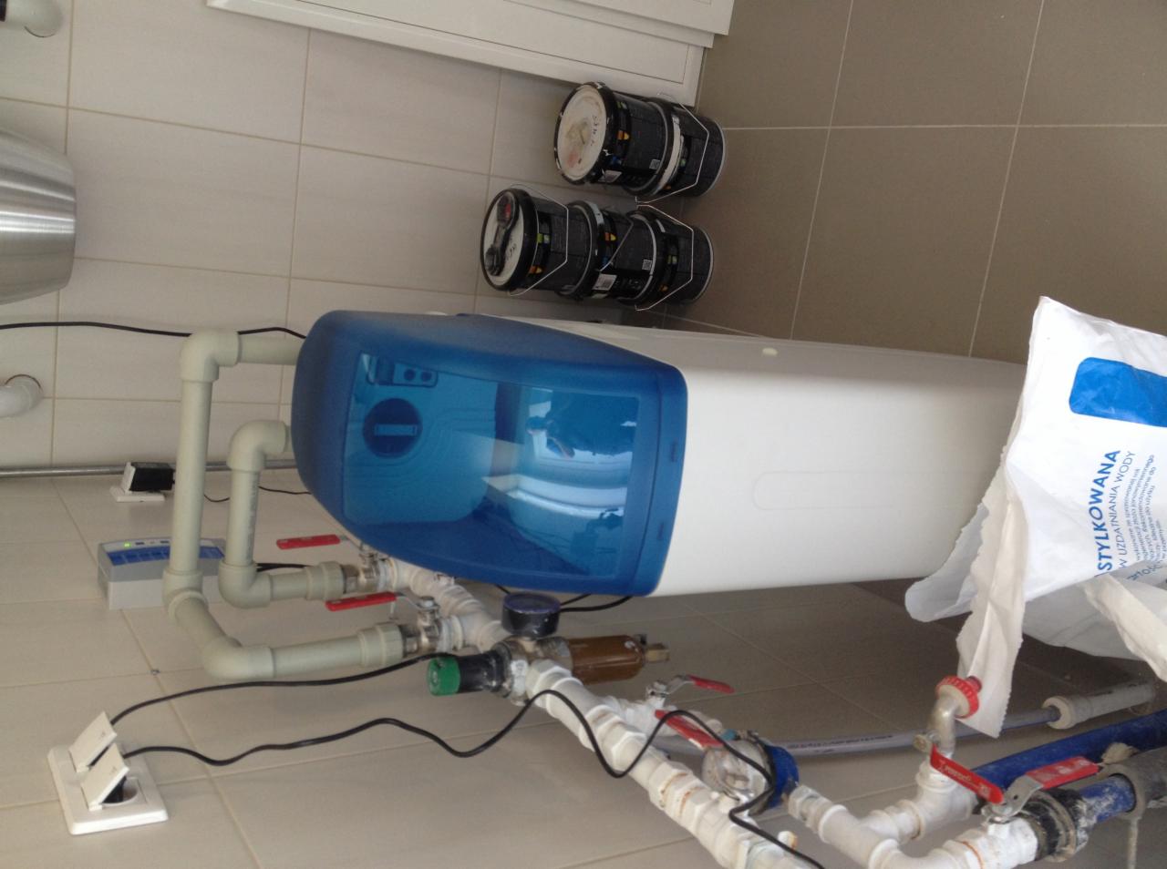 Biało-niebieski zmiękczacz wody z podłączonymi rurami instalacji wodnej, widoczne także opakowania soli do uzdatniania wody i puszki z farbą w tle.