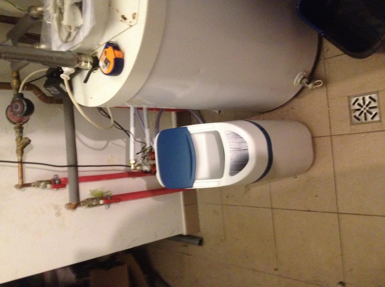 Biały zmiękczacz wody z niebieską pokrywą, zamontowany obok bojlera, widoczne rury instalacyjne z zaworami i wodomierz. Ujęcie z góry, na tle płytek podłogowych.