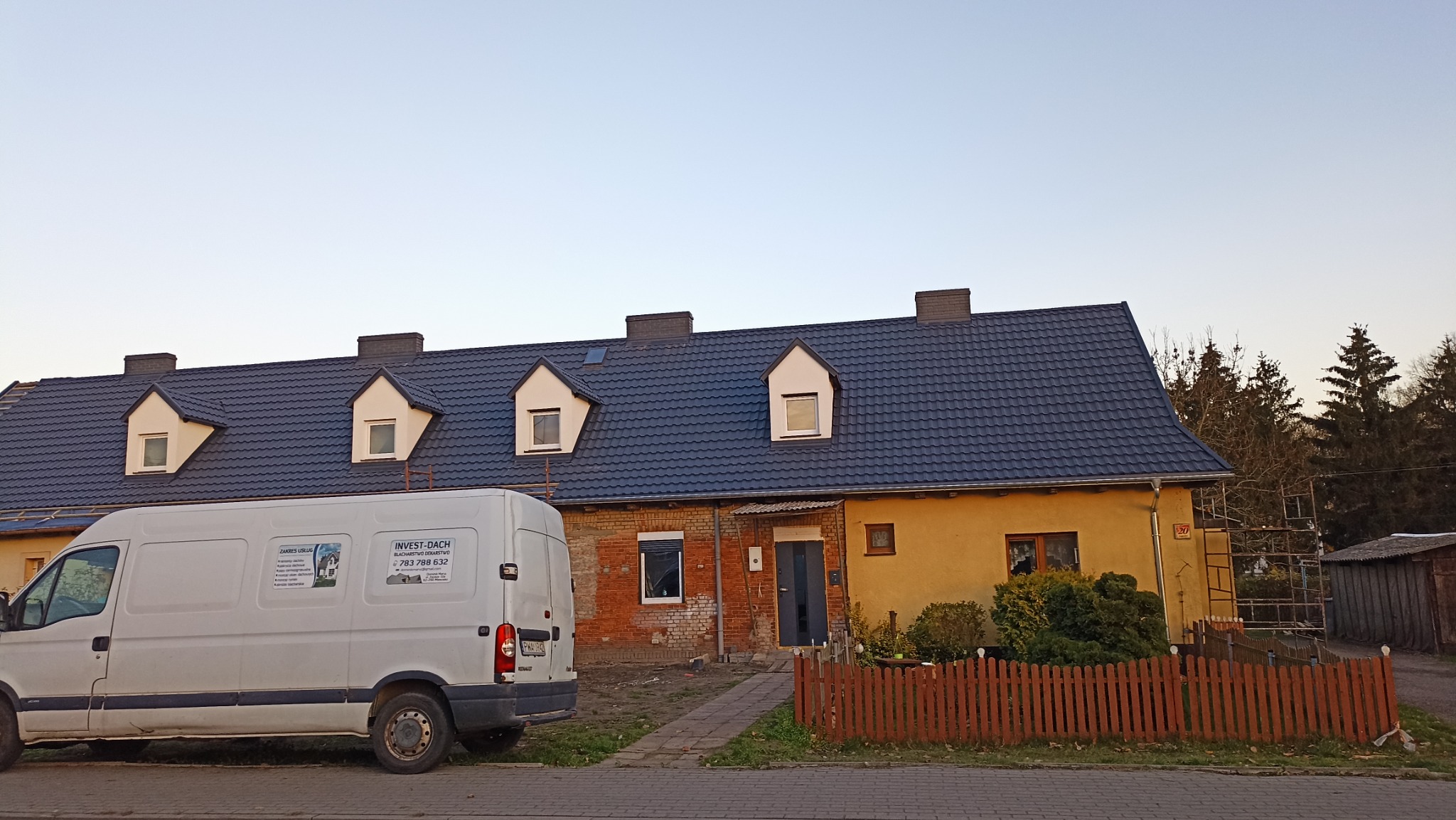 Dom po remoncie dachu z nową, ciemną dachówką, widoczny biały bus firmy dekarskiej z reklamą na boku i rusztowanie przy ścianie budynku.