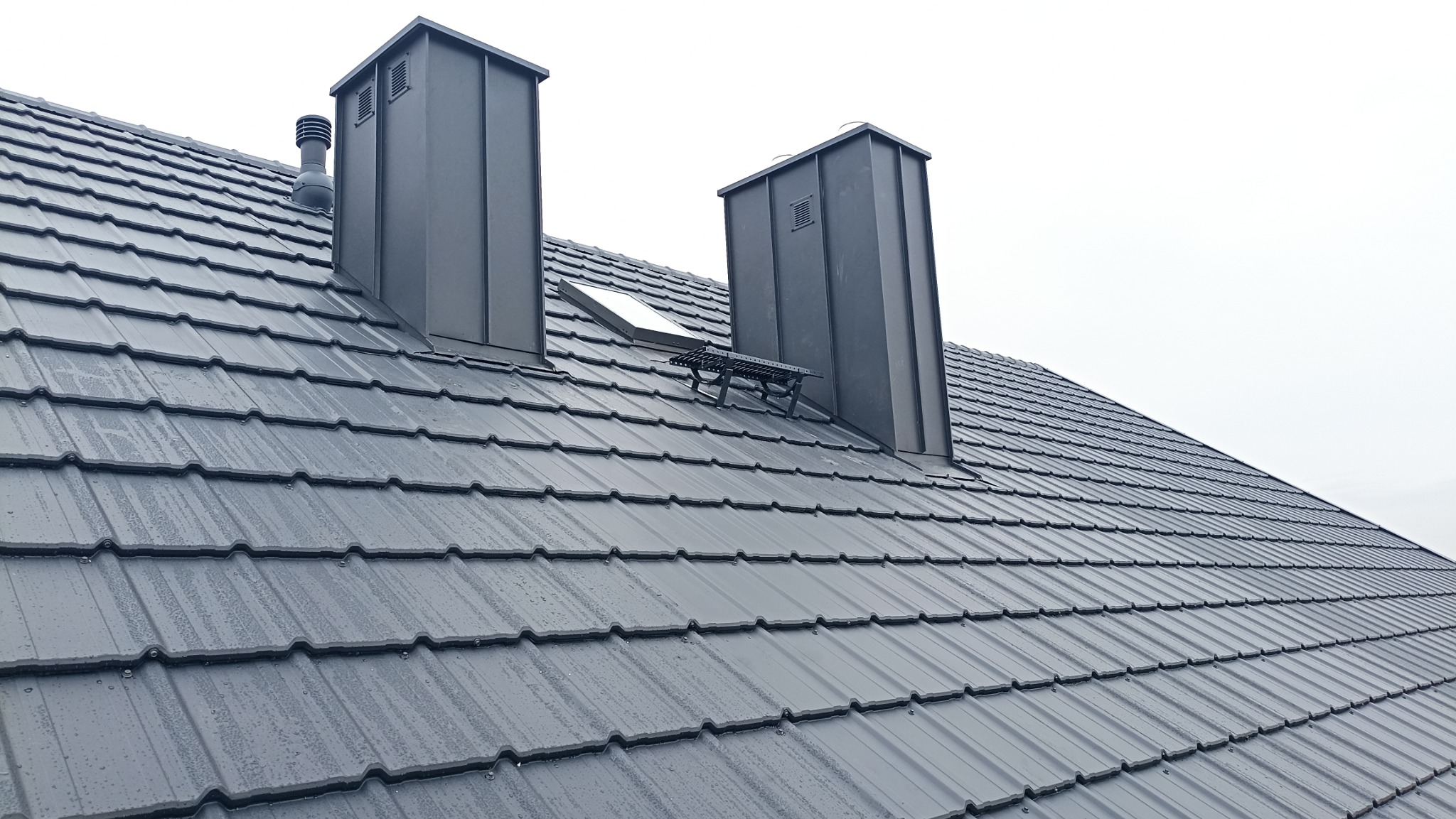 Szary dach z blachy trapezowej z dwoma kominami z blachy, okno dachowe i drabinka kominiarska, lekko wilgotny.