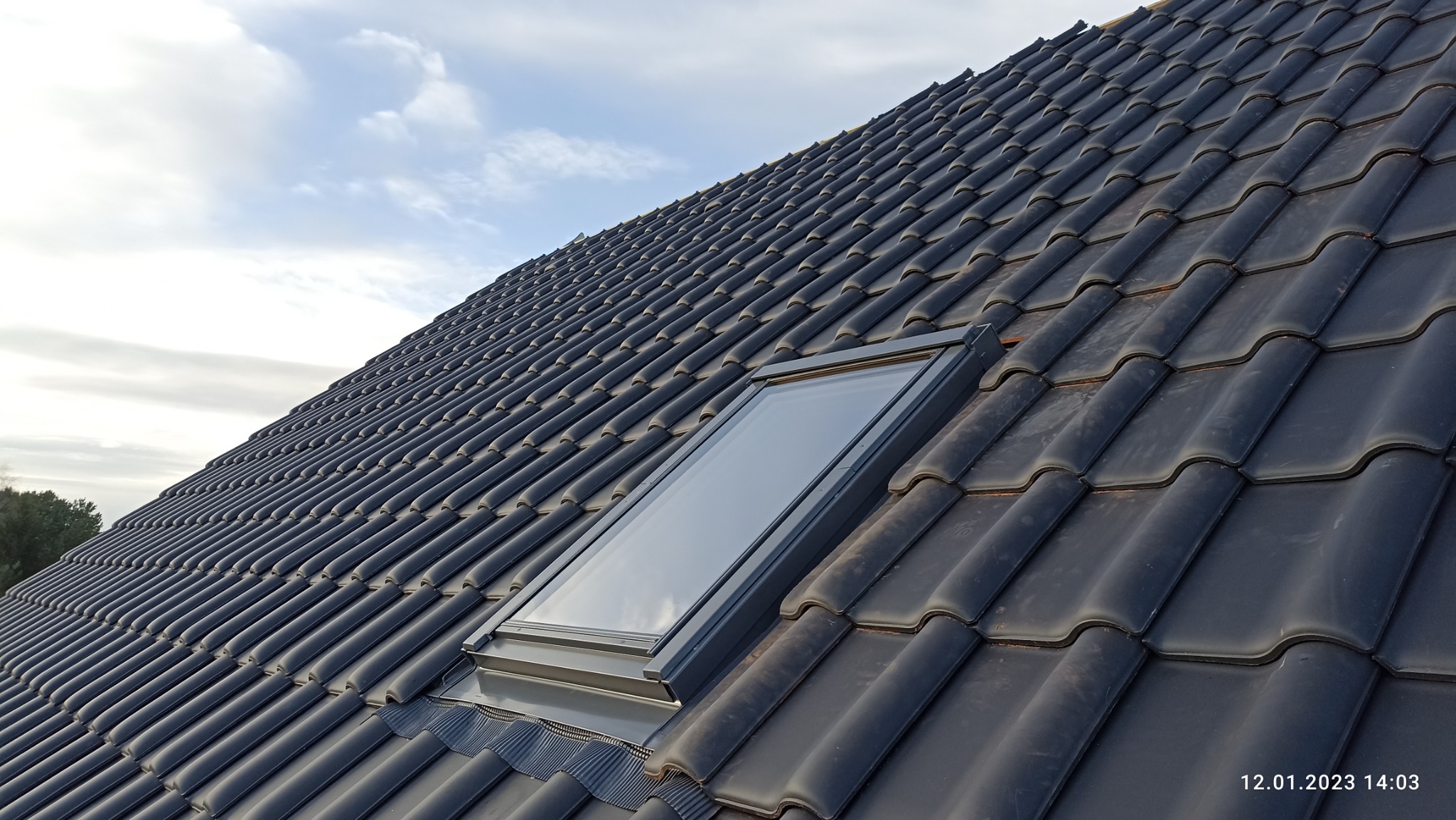 Nowy dach z czarnej dachówki ceramicznej z zamontowanym oknem dachowym, widok pod kątem, niebo z chmurami w tle.