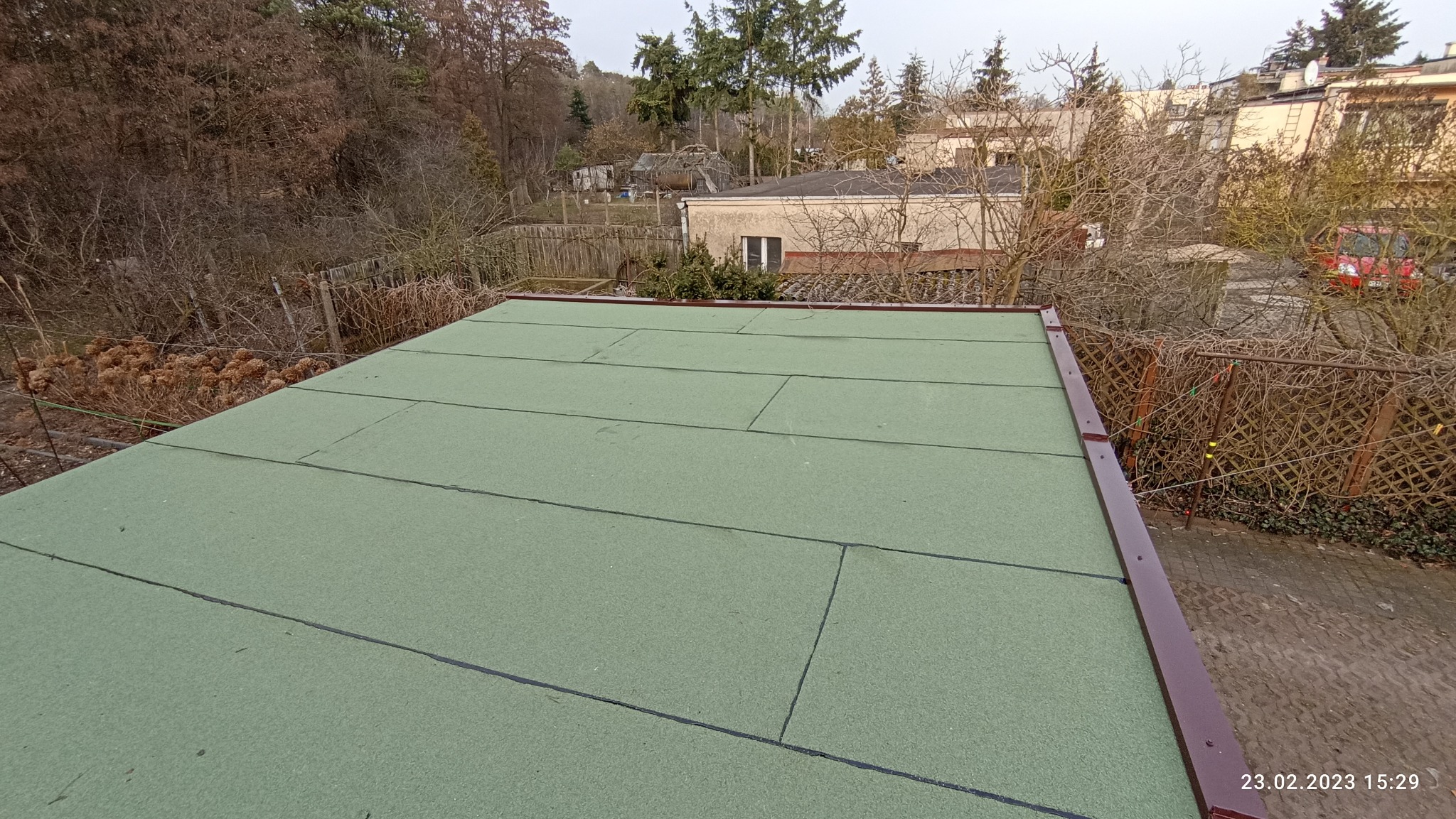 Widok z góry na płaski dach pokryty zieloną papą bitumiczną, z widocznymi łączeniami i metalową obróbką krawędzi w kolorze brązowym, na tle drzew i zabudowań.