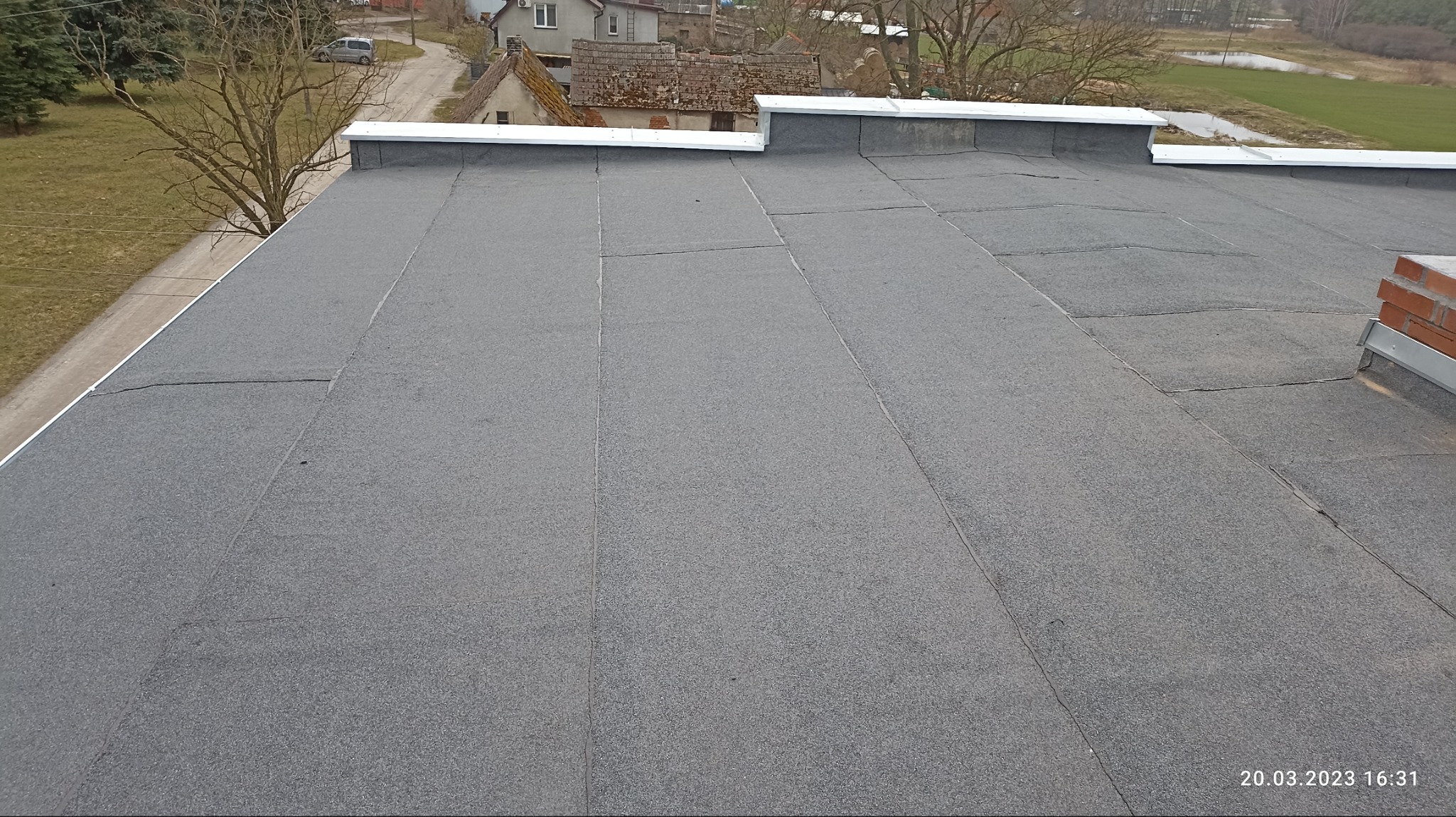 Szary, płaski dach pokryty papą termozgrzewalną, widoczne łączenia i komin z cegły w prawym rogu, w tle zabudowa wiejska i drzewa bez liści.