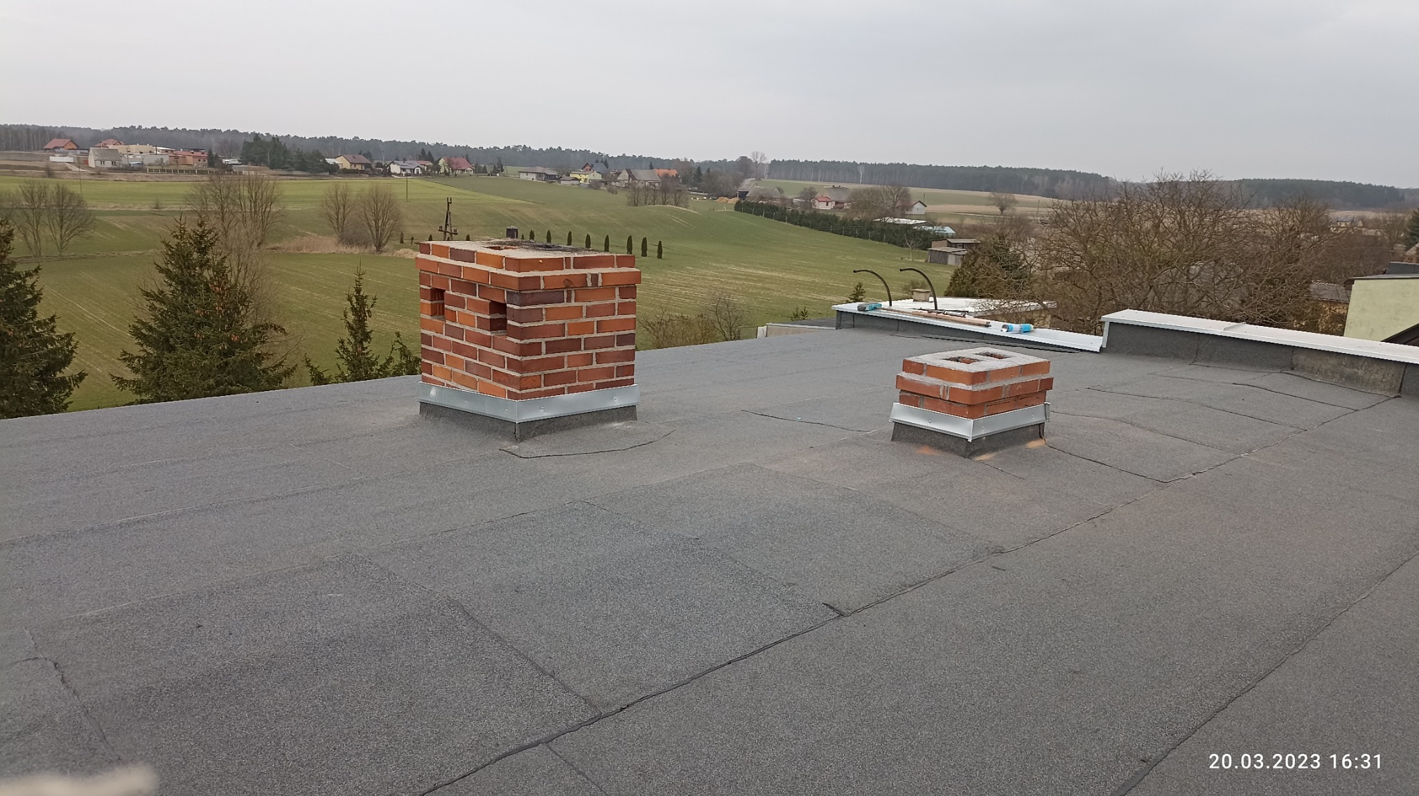 Dach pokryty papą termozgrzewalną z dwoma wymurowanymi kominami z cegły klinkierowej, widok na okoliczne pola i zabudowania.