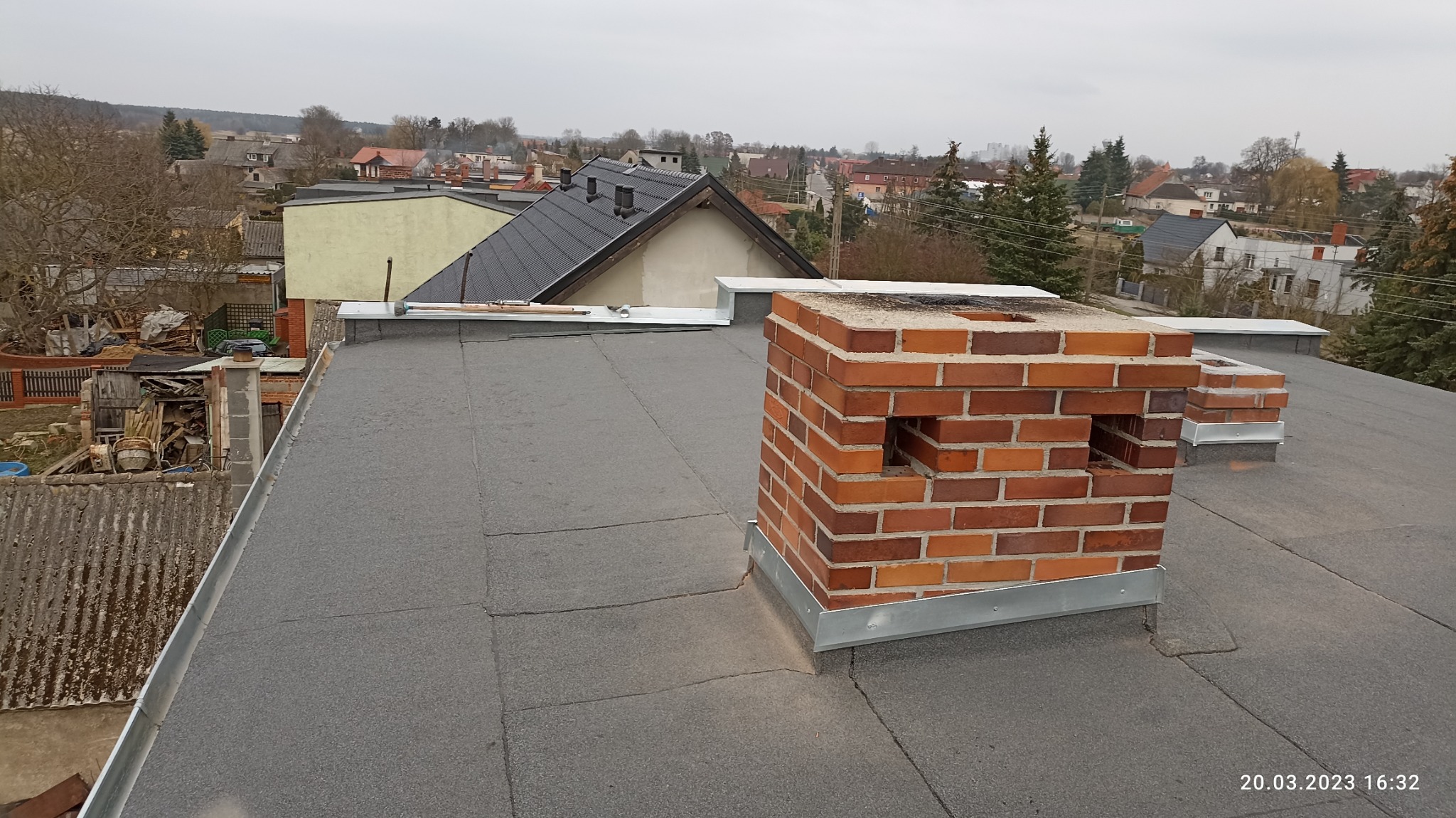 Widok z góry na płaski dach pokryty papą z ceglanym kominem z otworami wentylacyjnymi, w tle zabudowa jednorodzinna i pochmurne niebo.