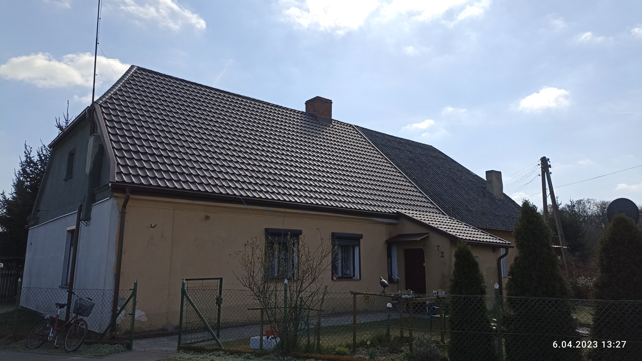 Dom jednorodzinny z nowym, brązowym dachem z blachodachówki, widoczne rynny, komin z cegły i fragment starego dachu z szarej dachówki bitumicznej po prawej stronie.