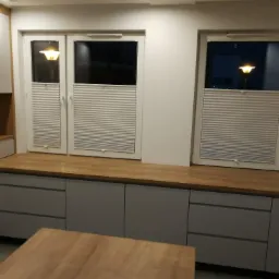 Wnętrze kuchni z szarymi szafkami i drewnianym blatem, widoczne trzy okna z zamontowanymi, częściowo zasłoniętymi plisowanymi roletami w kolorze białym.