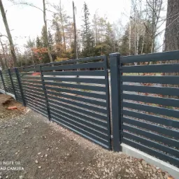 Długa, pozioma, metalowa konstrukcja ogrodzenia z szarych paneli, z widocznymi elementami mocującymi, osadzona na betonowej podmurówce i żwirowym podłożu, w tle drzewa bez liści.