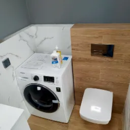 Mała łazienka z pralką Samsung, wiszącą toaletą i marmurowymi płytkami na ścianach, uzupełniona butelką płynu do prania na pralce.