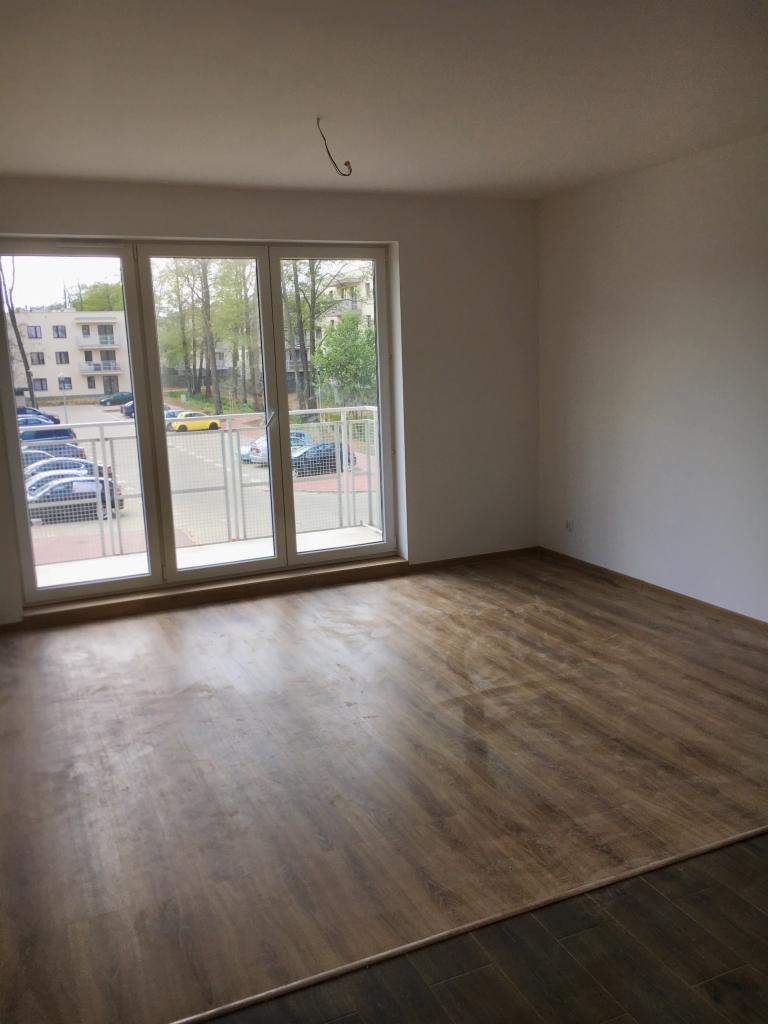 Pokój z nową podłogą z paneli imitujących drewno, widok na balkon i osiedle przez duże okno balkonowe, surowe ściany przygotowane do malowania.