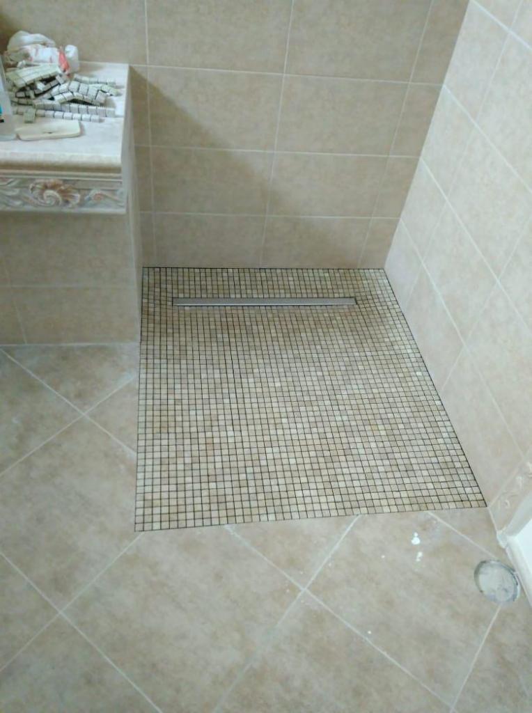 Wykończony brodzik z mozaiki w beżowych odcieniach, z odpływem liniowym, sąsiadujący z płytkami podłogowymi o większym formacie i blatem z resztkami płytek mozaikowych.