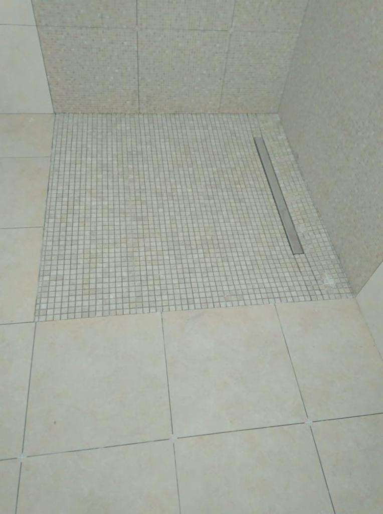 Wyłożony mozaiką brodzik z odpływem liniowym w łazience w Warszawie, otoczony beżowymi płytkami podłogowymi i ściennymi.