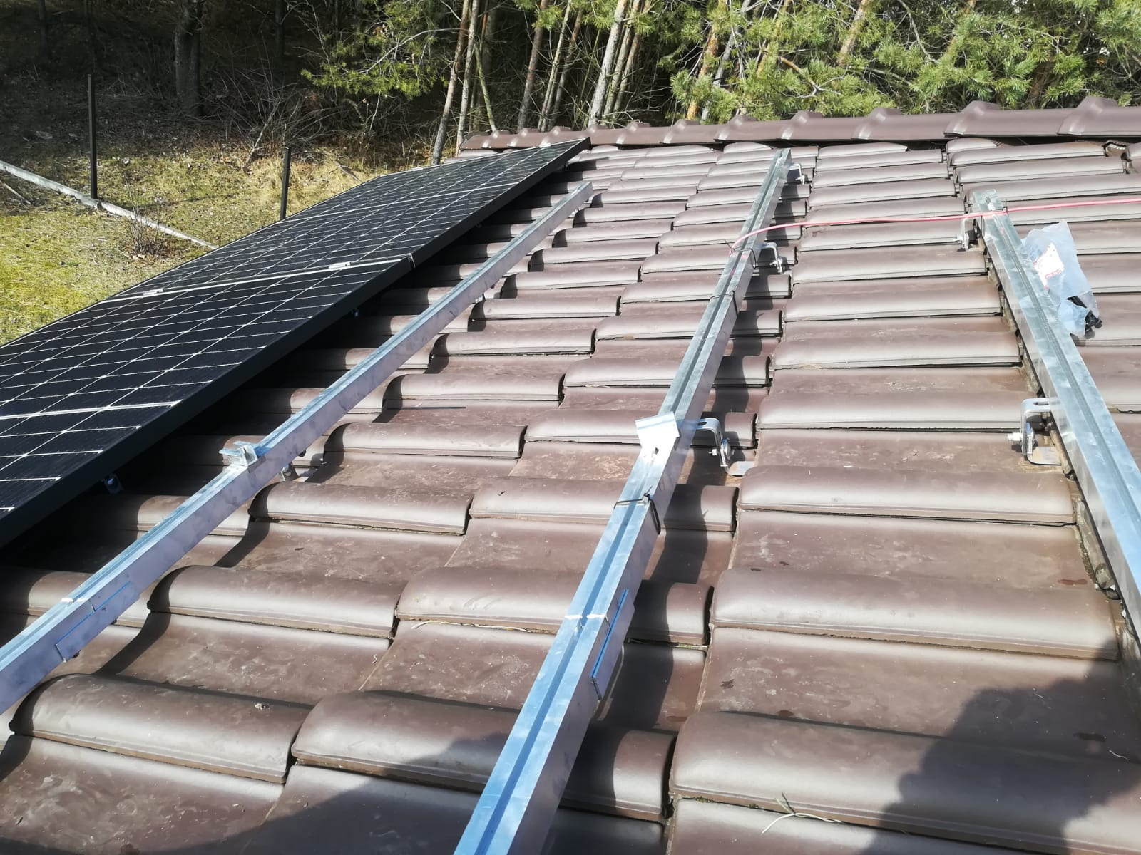 Częściowo zamontowane panele fotowoltaiczne na brązowym, skośnym dachu z dachówki ceramicznej, widoczne aluminiowe szyny montażowe i elementy mocujące.