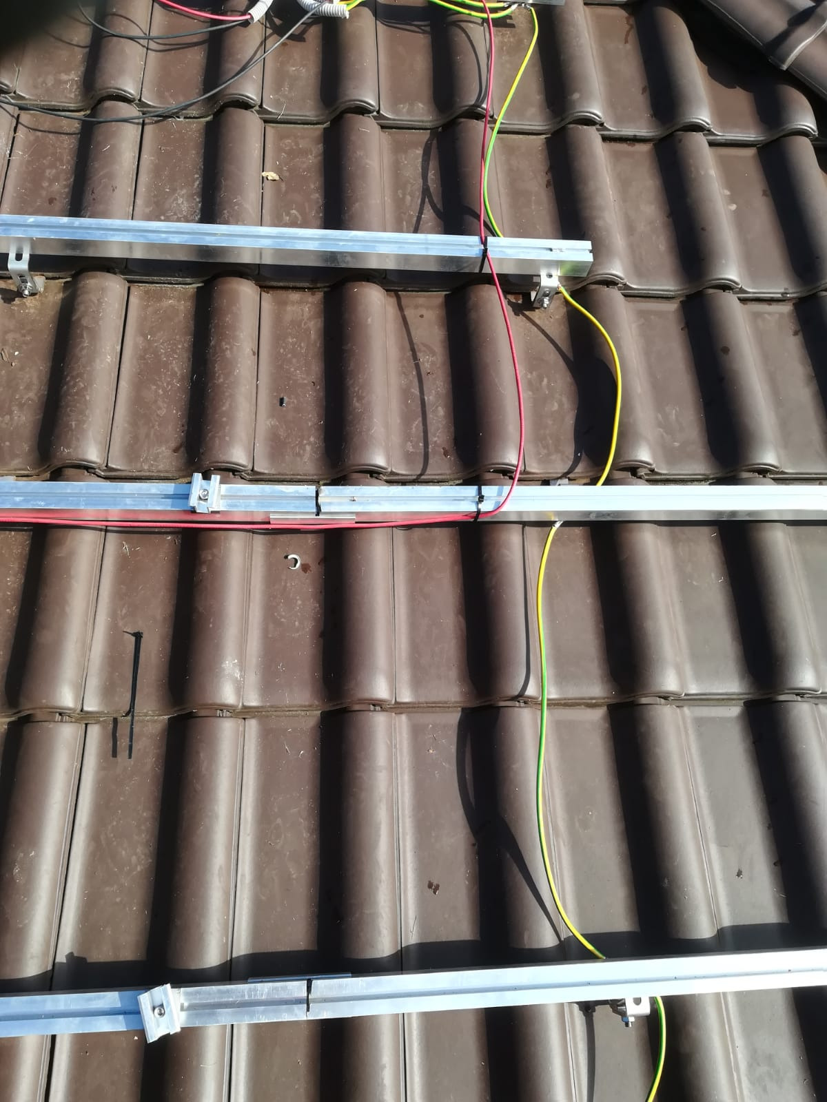 Montaż aluminiowych profili na brązowej dachówce pod instalację paneli słonecznych, widoczne przewody elektryczne w kolorach czerwonym, żółtym i zielonym.