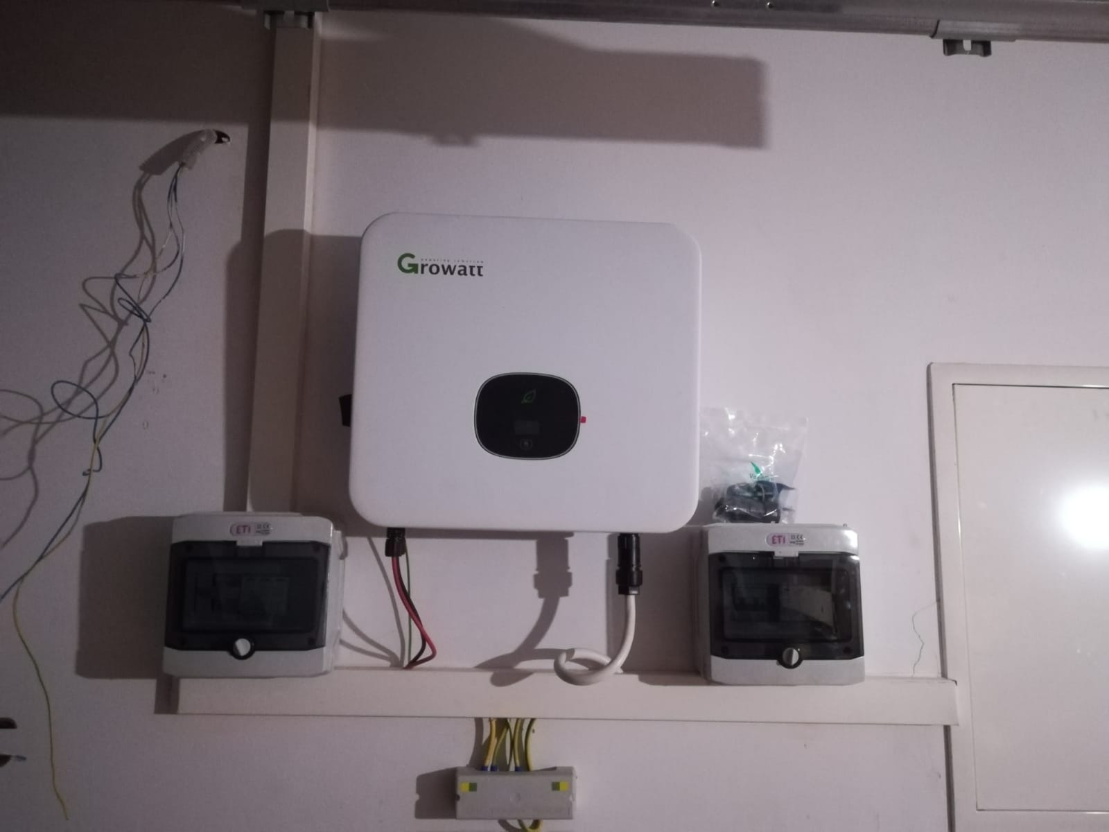 Biały inwerter Growatt zamontowany na ścianie, po bokach dwie szare skrzynki elektryczne, widoczne przewody i kable w ścianie.