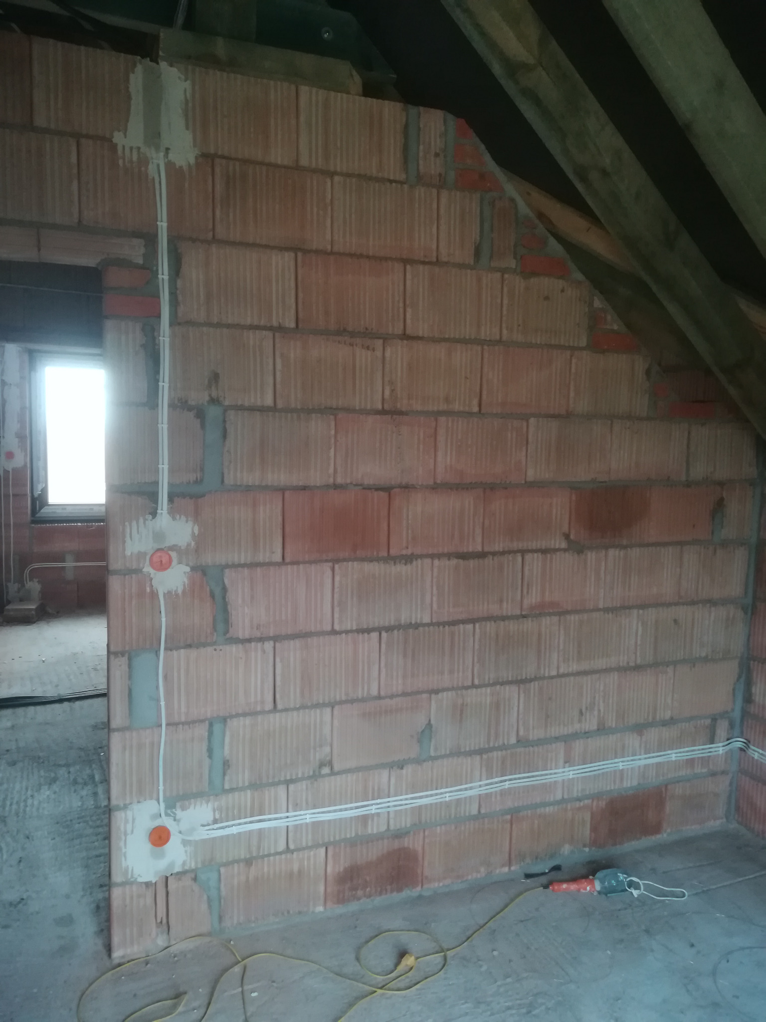 Surowa ściana z czerwonej cegły w trakcie kładzenia instalacji elektrycznej, widoczne puszki elektryczne i przewody poprowadzone po ścianie, na podłodze leży wiertarka