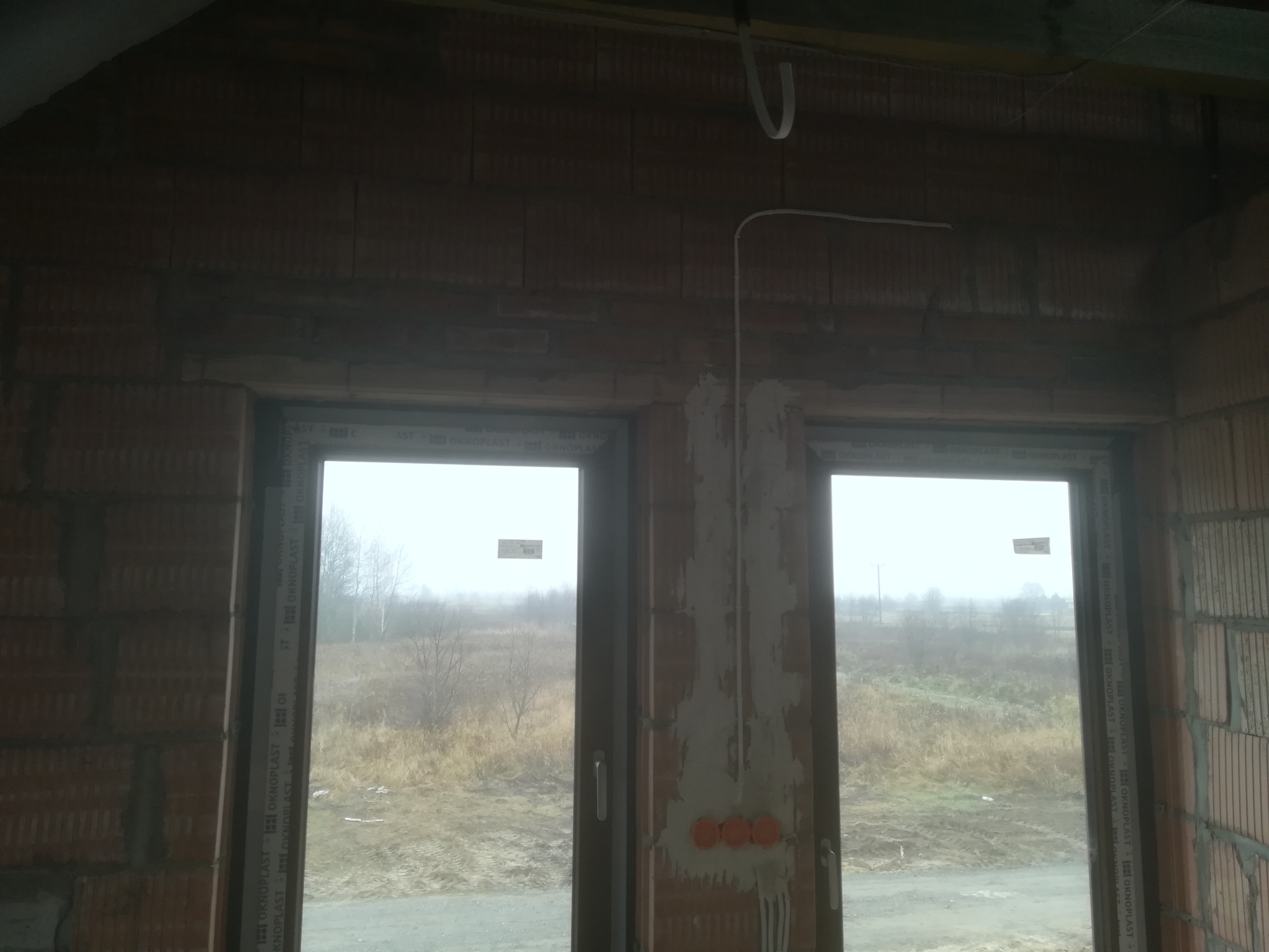 Wnętrze surowego budynku z widocznymi dwoma oknami, instalacją elektryczną w ścianie pomiędzy oknami oraz przewodami elektrycznymi pod sufitem. Widoczne puszki elektryczne w ścianie. Na zewnątrz...