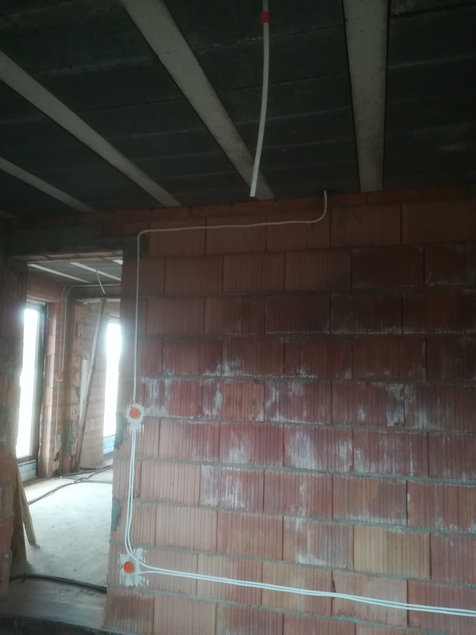 Surowy mur z cegły z poprowadzonymi przewodami elektrycznymi w białych rurkach, widoczne puszki instalacyjne pomarańczowe, sufit betonowy z widocznymi belkami konstrukcyjnymi, stan surowy budynku.