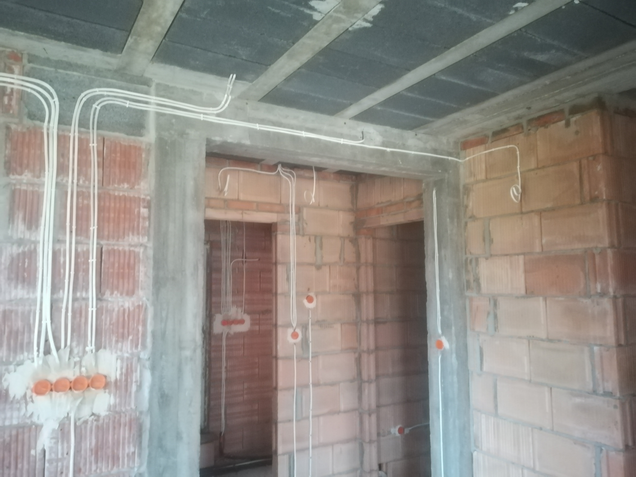 Surowy stan budynku w trakcie prac instalacyjnych: widoczne przewody elektryczne prowadzone po ceglanych ścianach i betonowym suficie, z puszkami instalacyjnymi w kolorze pomarańczowym.