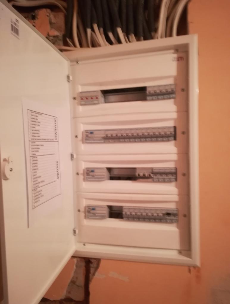 Otwarta skrzynka elektryczna z widocznymi bezpiecznikami i listwą zaciskową, zamontowana na pomarańczowej ścianie, z wystającymi kablami u góry i kartką z opisem obwodów na wewnętrznej stronie...