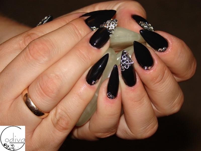 Dłonie z czarnymi, błyszczącymi paznokciami w kształcie migdała i szpicu, ozdobionymi srebrnymi cyrkoniami, trzymające jasne kamienie; na palcu złoty pierścionek.