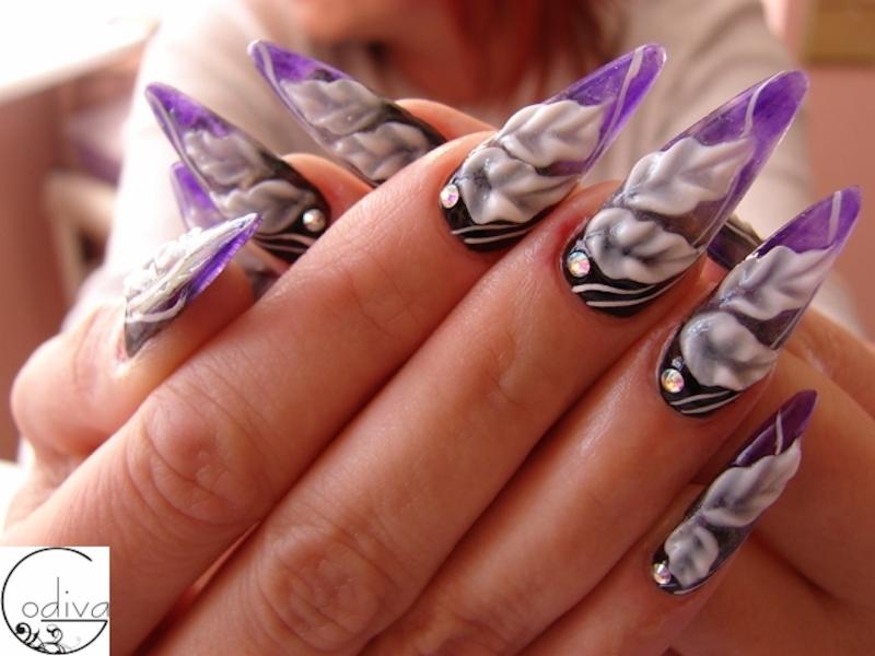 Dłoń z długimi, fioletowo-białymi paznokciami o szpiczastym kształcie, zdobionymi trójwymiarowym wzorem kwiatowym i drobnymi kryształkami u nasady.