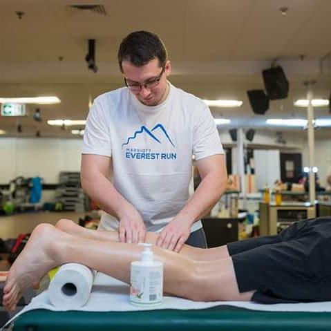 Fizjoterapeuta masuje łydki pacjenta leżącego na zielonym stole do masażu, widoczna butelka balsamu i rolka papieru.