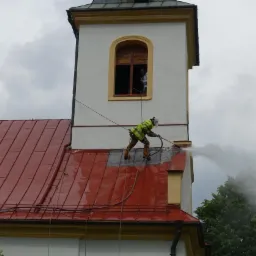 Pracownik w kombinezonie ochronnym i masce, zabezpieczony linami, czyści ciśnieniowo fragment czerwonego dachu kościoła, z wieżą w tle, widoczna osoba w oknie wieży.