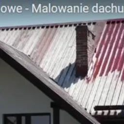 Dwóch robotników w białych kombinezonach, zabezpieczonych linami, maluje blaszany dach na czerwono. Część dachu jest już pomalowana, a część jeszcze w kolorze srebrnym. Widoczny komin i fragment domu.