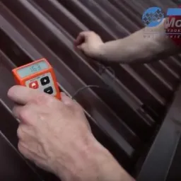 Mężczyzna mierzy wilgotność brązowego dachu z blachy trapezowej za pomocą wilgotnościomierza elektronicznego, widoczne logo firmy na dachu.