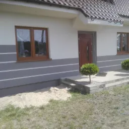 Wykończona elewacja domu jednorodzinnego z szarymi pasami, brązowymi oknami i drzwiami wejściowymi; widoczne dwa małe, kuliste drzewka przed wejściem i piasek przy fundamencie.