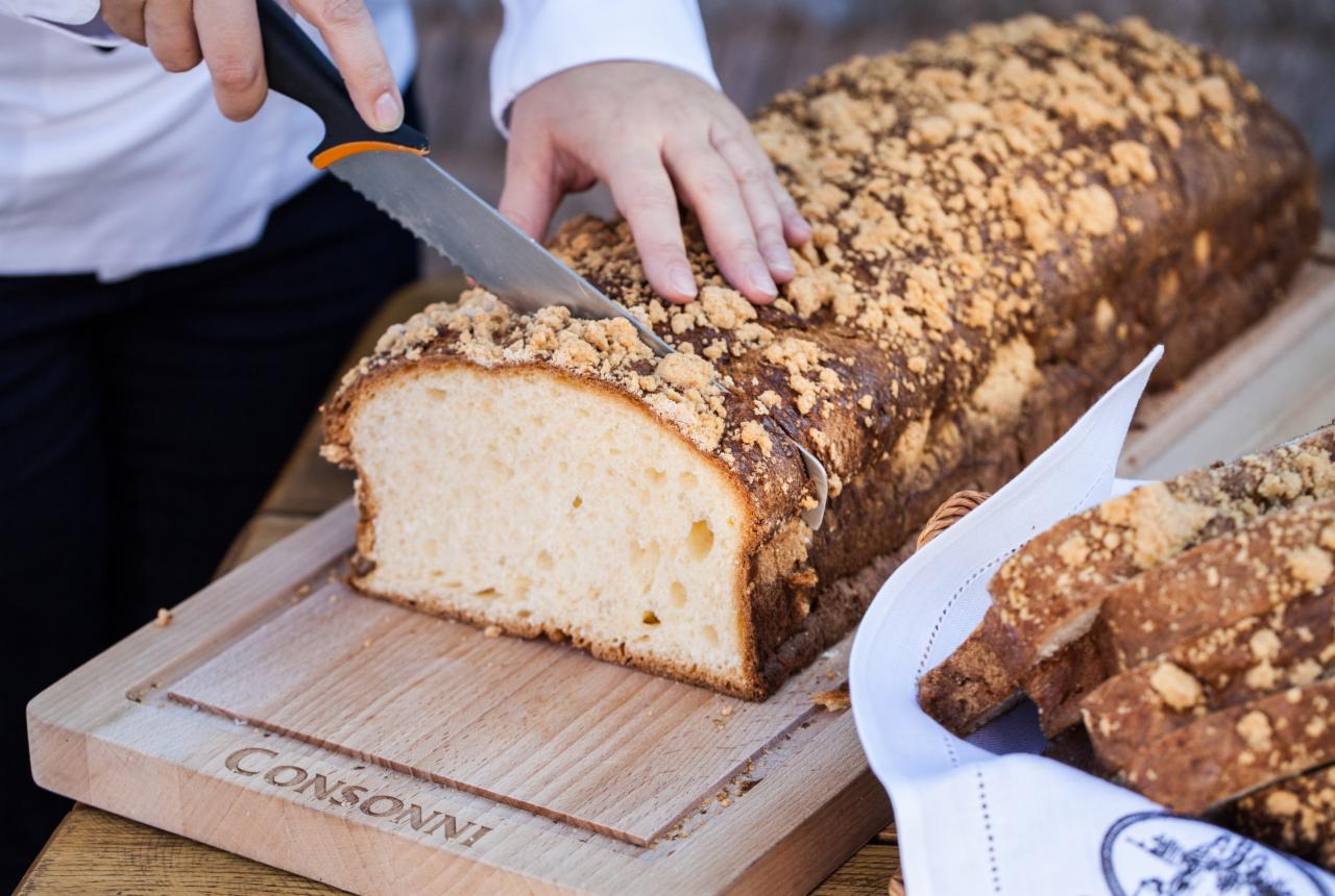 Krojenie babki drożdżowej z kruszonką na drewnianej desce z wygrawerowanym napisem 'Consonni', obok koszyk z pokrojonymi kromkami.