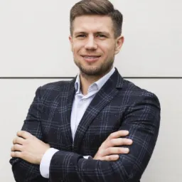 Uśmiechnięty mężczyzna w garniturze w kratę pozuje na tle jasnej ściany, prezentując profesjonalny wygląd.