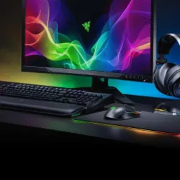 Gamingowy zestaw komputerowy Razer: monitor z tęczowym tłem, podświetlana klawiatura z podkładką pod nadgarstki, mysz i podkładka z obwódką RGB oraz słuchawki na stojaku, wszystko na czarnym biurku...