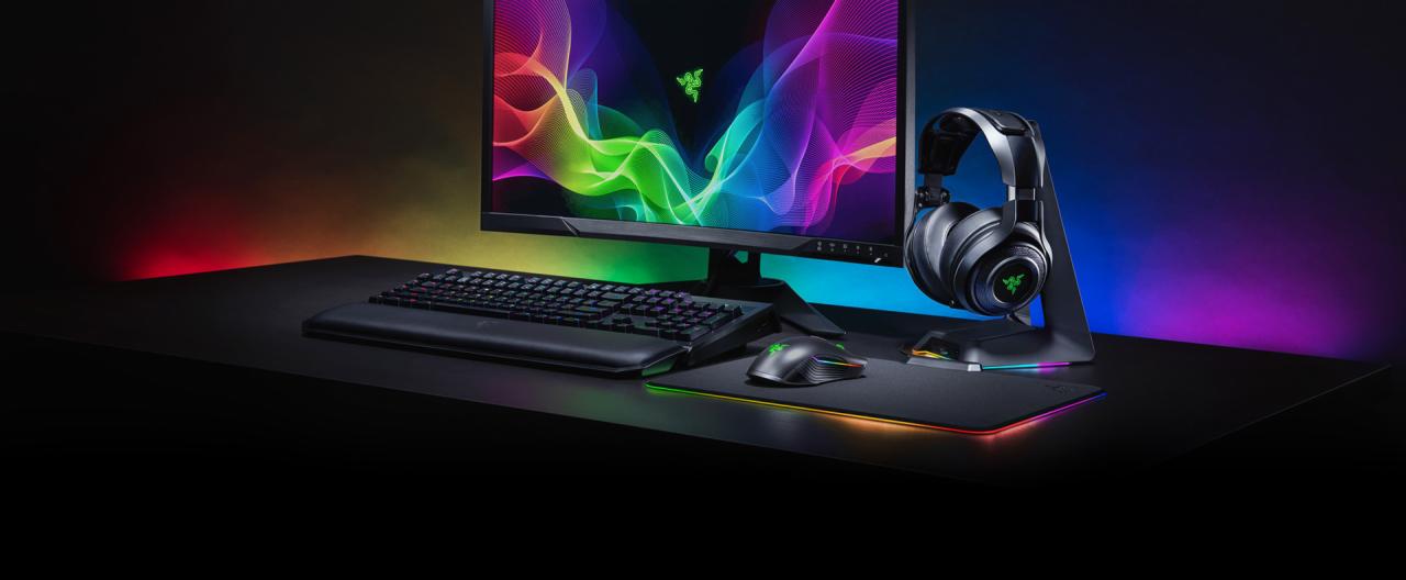 Gamingowy zestaw komputerowy Razer: monitor z tęczowym tłem, podświetlana klawiatura z podkładką pod nadgarstki, mysz i podkładka z obwódką RGB oraz słuchawki na stojaku, wszystko na czarnym biurku...