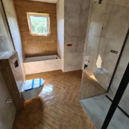 Nowoczesna łazienka w trakcie remontu: brązowa podłoga ułożona w jodełkę, częściowo wykończona kabina prysznicowa z czarnymi profilami, zabezpieczona toaleta i fragment wanny pod oknem.