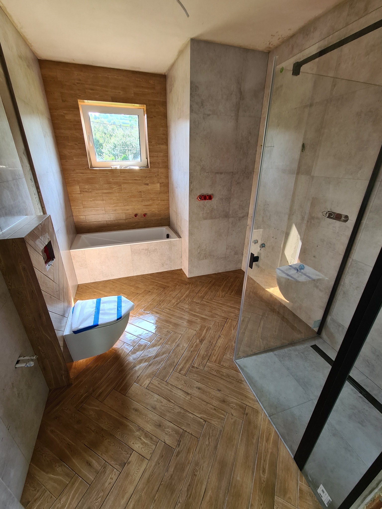 Nowoczesna łazienka w trakcie remontu: brązowa podłoga ułożona w jodełkę, częściowo wykończona kabina prysznicowa z czarnymi profilami, zabezpieczona toaleta i fragment wanny pod oknem.