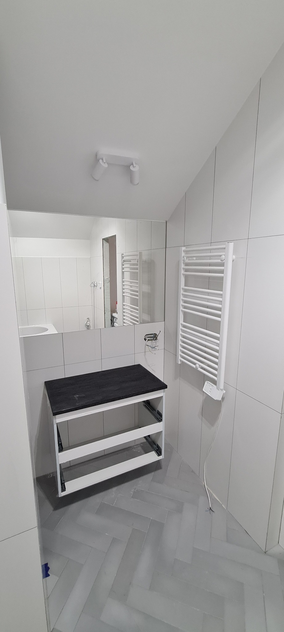 Łazienka w trakcie wykańczania: widoczna szafka pod umywalkę z czarnym blatem bez frontów szuflad, biały grzejnik drabinkowy z podłączonym kablem, lustro i szare płytki ułożone w jodełkę na podłodze.