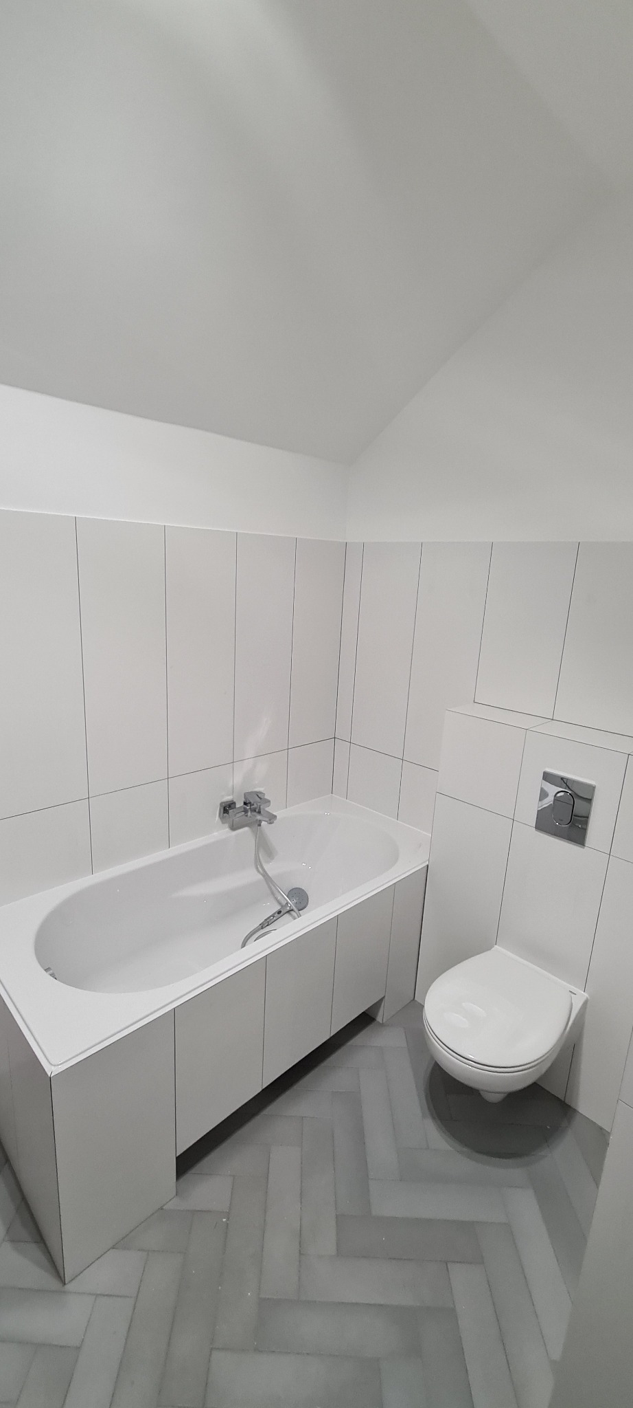 Nowoczesna łazienka w bieli i szarości: wanna obudowana panelami, podwieszana toaleta z chromowanym przyciskiem, szare płytki ułożone w jodełkę na podłodze, pionowe białe płytki na ścianach.