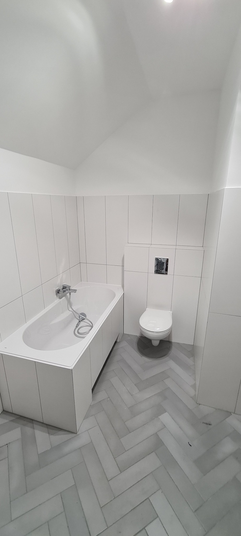 Biała łazienka z wanną, podwieszanym WC i szarą podłogą ułożoną w jodełkę, wykończona białymi, pionowymi płytkami.