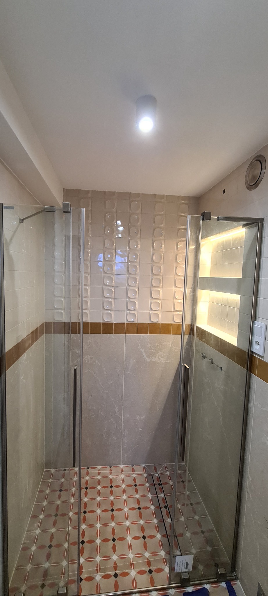 Nowoczesna kabina prysznicowa z szklanymi drzwiami, wykończona beżowymi płytkami z dekoracyjnym pasem i wzorzystą podłogą w odcieniach czerwieni i beżu.