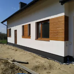 Nowoczesny dom murowany z ocieploną elewacją, wykończoną panelami imitującymi drewno wokół okien i drzwi, widoczne prace wykończeniowe wokół budynku.