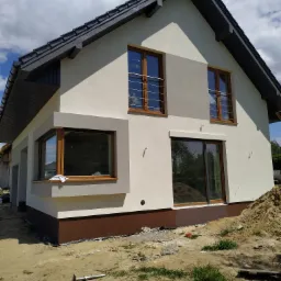 Nowoczesny dom jednorodzinny z elewacją w odcieniach bieli i szarości, z brązowymi ramami okiennymi i ciemnoszarą dachówką, w trakcie budowy, z widocznymi elementami rusztowania i stertą ziemi...