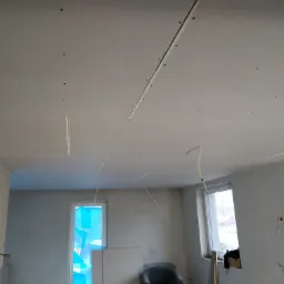 Wnętrze w trakcie montażu sufitu z płyt kartonowo-gipsowych, widoczne profile i otwory na wkręty, okna zabezpieczone folią malarską, wystające przewody elektryczne.