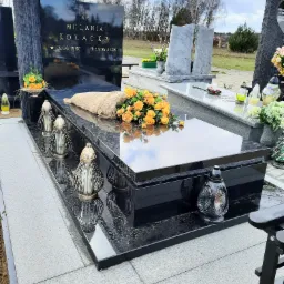 Czarny, polerowany nagrobek z inskrypcją 'Ś.P. MELANIA KOŁACKA' i datami urodzenia i śmierci, ozdobiony bukietem żółtych róż i metalowymi lampionami, na tle innych nagrobków i drzew.
