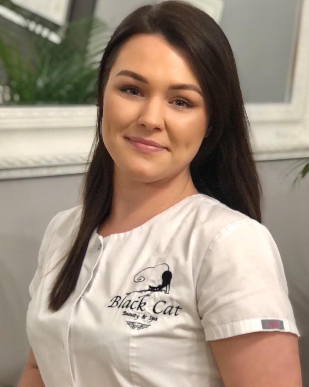 Portret uśmiechniętej brunetki w białym uniformie z logo Black Cat Beauty & Spa, stojącej w salonie z roślinami w tle.