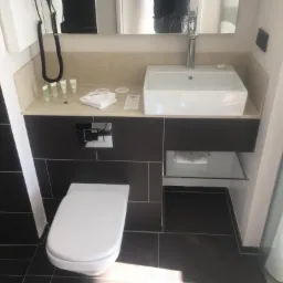 Nowoczesna łazienka z czarnymi płytkami, białą ceramiką i umywalką w kształcie prostopadłościanu, widok na toaletę i szklaną półkę pod umywalką.